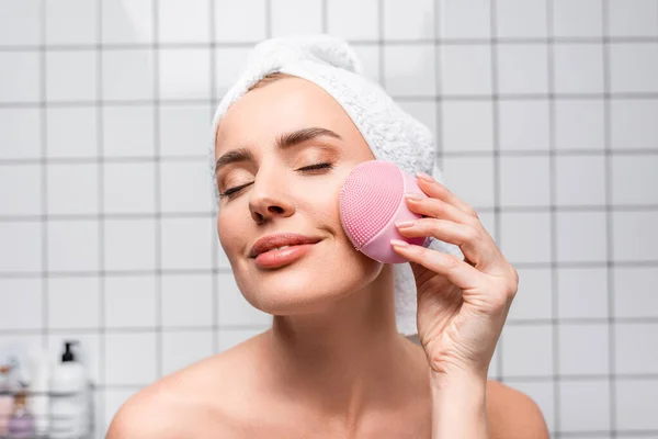 Cheerful woman in towel on head and closed eyes holding cleansing silicone brush in bathroom — Stock Photo