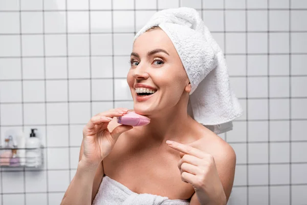 Donna allegra in asciugamano sulla testa che punta con il dito a spazzola di silicone detergente in bagno — Foto stock
