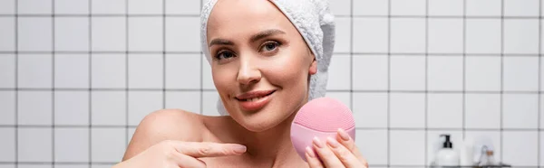 Cheerful woman in towel on head pointing with finger at cleansing silicone brush in bathroom — Stock Photo