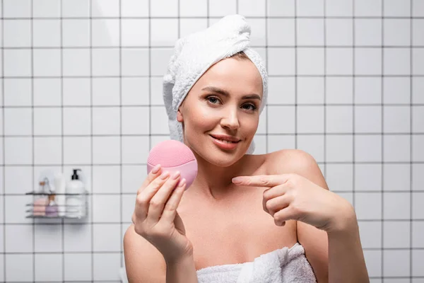 Joyeuse femme en serviette sur la tête pointant du doigt à la brosse en silicone dans la salle de bain — Photo de stock