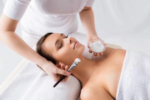 Beautician holding cosmetic brush and container with face mask near client with closed eyes lying on massage table — Stock Photo