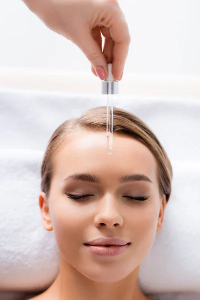 Draufsicht der Kosmetikerin, die Pipette hält und Serum auf das Gesicht der lächelnden Frau im Wellness-Salon aufträgt — Stockfoto