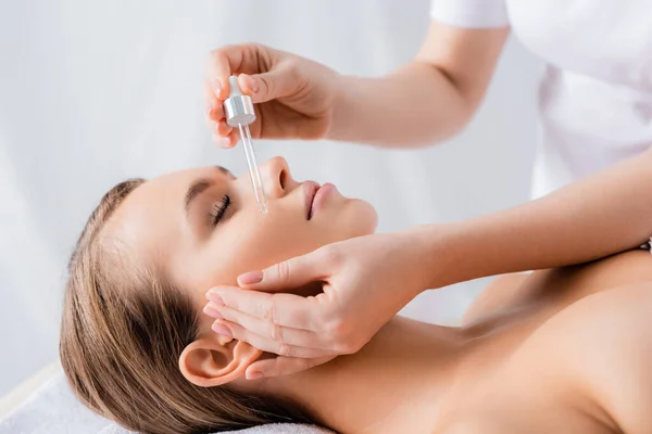 Esteticista sosteniendo la pipeta y aplicando suero en la cara de la mujer con los ojos cerrados en el salón de spa — Stock Photo