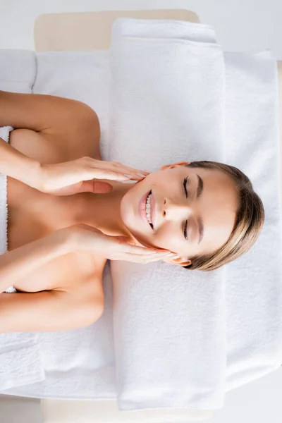 Vista superior da mulher feliz tocando bochechas e deitado na mesa de massagem no salão de spa — Fotografia de Stock