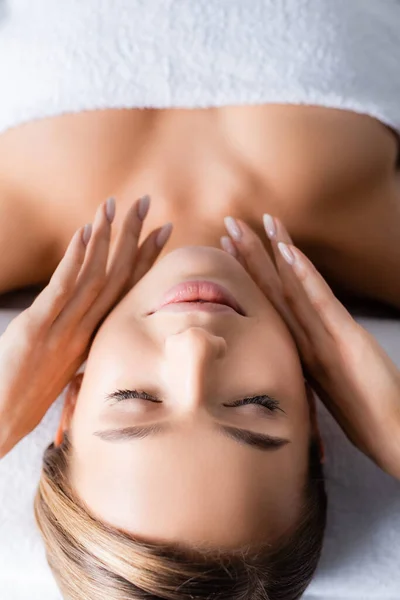 Mujer joven con los ojos cerrados tocando la cara y acostado en la mesa de masaje en el salón de spa - foto de stock