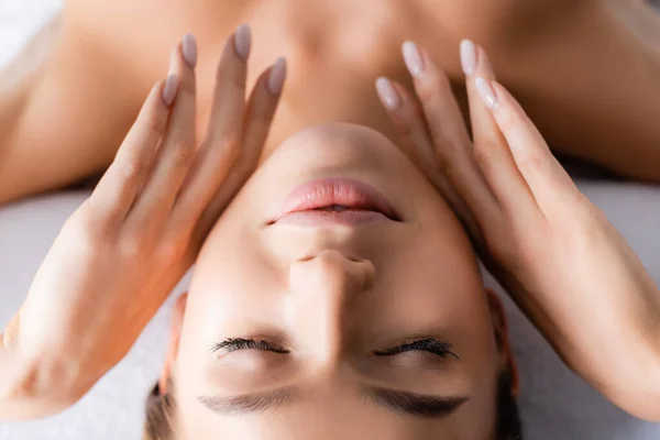 Jeune femme aux yeux fermés touchant le visage et allongée sur une table de massage dans un salon de spa — Photo de stock