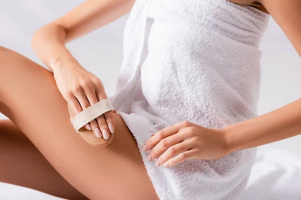 Cropped view of young woman wrapped in white towel exfoliating skin on leg — Stock Photo