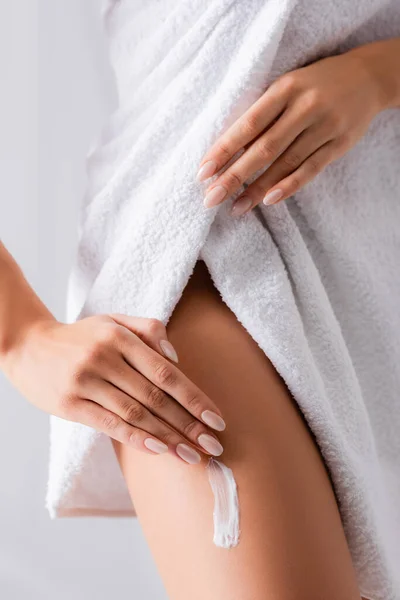 Vista parcial de mujer joven envuelta en toalla aplicando crema cosmética en la pierna sobre blanco - foto de stock