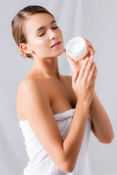 Mujer joven con los ojos cerrados y crema en la cara sosteniendo frasco en blanco - foto de stock