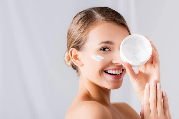 Alegre joven con crema en la cara sosteniendo tarro y mirando a la cámara en blanco — Stock Photo