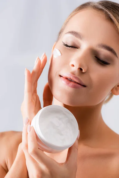 Mujer con hombros desnudos sosteniendo el frasco y aplicando crema facial sobre blanco — Stock Photo