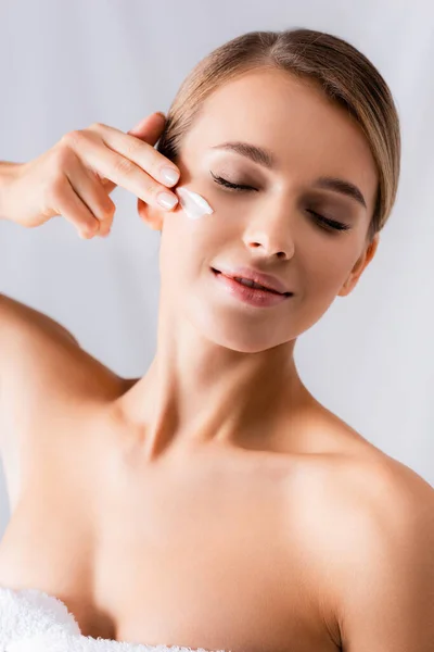 Cheerful young woman with bare shoulders applying face cream on white — Stock Photo