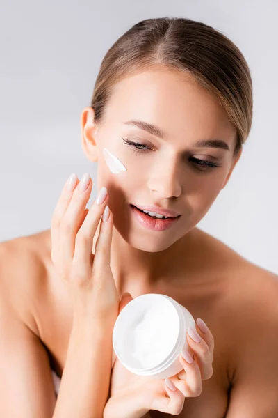 Mujer joven con hombros desnudos sosteniendo el frasco y aplicando crema facial aislada en blanco - foto de stock