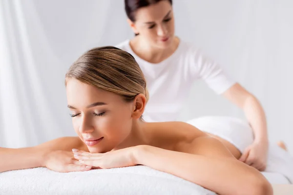 Jovem mulher deitada na mesa de massagem perto de massagista no fundo borrado — Fotografia de Stock