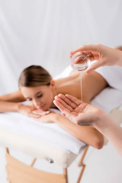 Masajista sosteniendo contenedor de vidrio y vertiendo aceite en la mano cerca de la mujer descansando sobre la mesa de masaje y fondo borroso - foto de stock