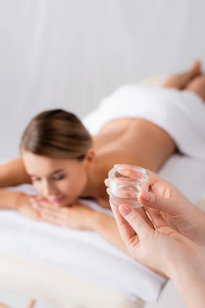 Massagista segurando recipiente de vidro com óleo perto do cliente descansando na mesa de massagem no fundo borrado — Fotografia de Stock
