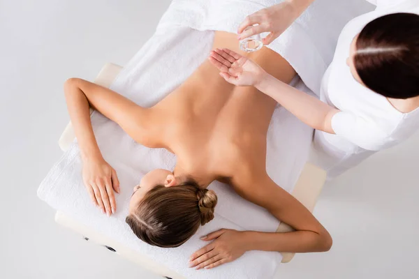 Top view masseur holding glass bottle and pouring massage oil on hand near woman in spa salon — Stock Photo