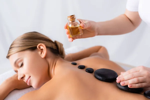 Masseur holding bottle with oil near client getting hot stone massage — Stock Photo