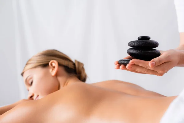 Massaggiatore in possesso di pietre calde vicino al cliente su sfondo sfocato — Foto stock