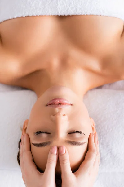 Vista superior de masajista haciendo masaje facial a mujer joven en salón de spa - foto de stock