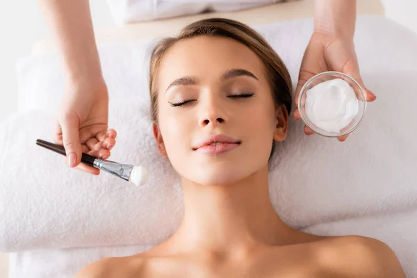 Vista superior de esteticista segurando escova cosmética e recipiente com máscara facial perto de jovem na mesa de massagem — Fotografia de Stock