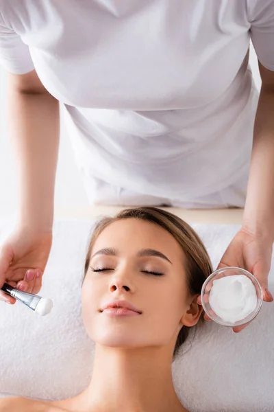 Esthéticienne tenant brosse cosmétique et récipient avec masque facial près de la jeune femme sur table de massage — Photo de stock