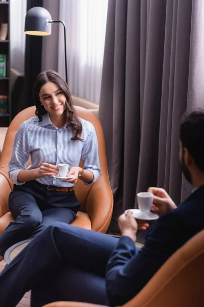 Sonriente mujer de negocios con taza de café cerca de árabe colega de negocios sentado en la zona de salón, borrosa primer plano - foto de stock
