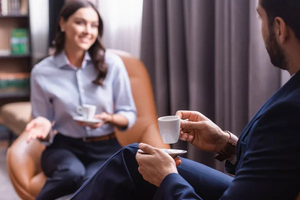 Musulmán hombre de negocios con taza de café cerca sonriente socio de negocios sentado en la zona de salón sobre fondo borroso - foto de stock