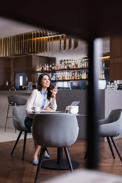 Happy freelancer looking away while sitting near laptop in restaurant, blurred front ground — стоковое фото