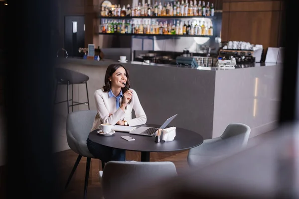 Счастливый, мечтательный фрилансер, отводящий взгляд, сидя возле ноутбука в ресторане — стоковое фото