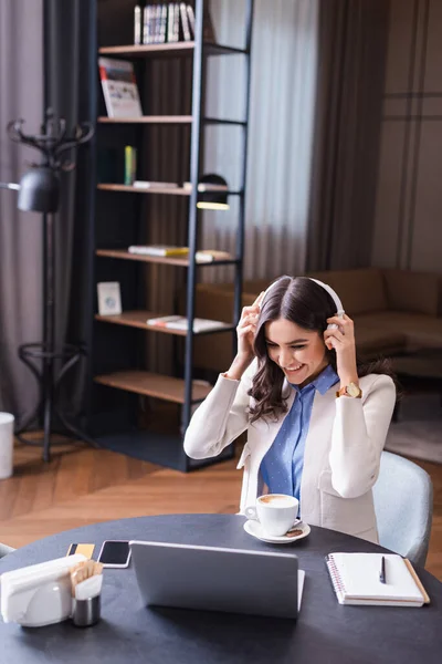 Cheerful freelancer putting on headphones while sitting in restaurant near laptop — Stock Photo