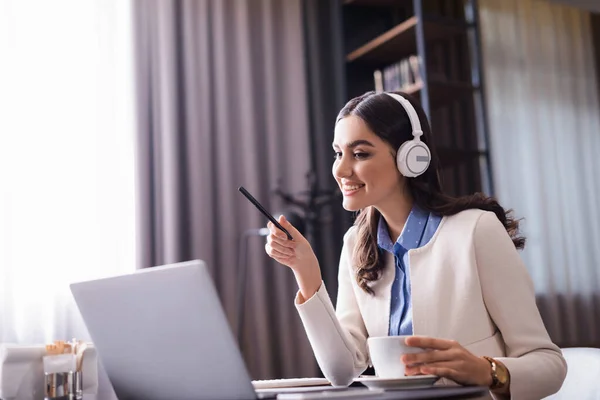 Cheerful freelancer in headphones pointing with pen at laptop while working in restaurant — Stock Photo