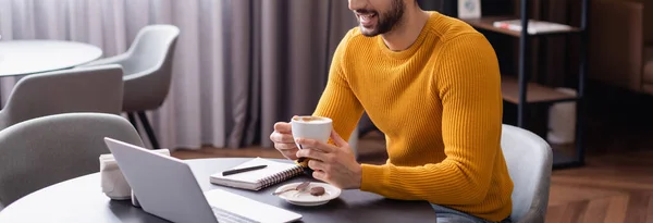 Vue recadrée de pigiste arabe tenant tasse de café près d'un ordinateur portable, bannière — Photo de stock