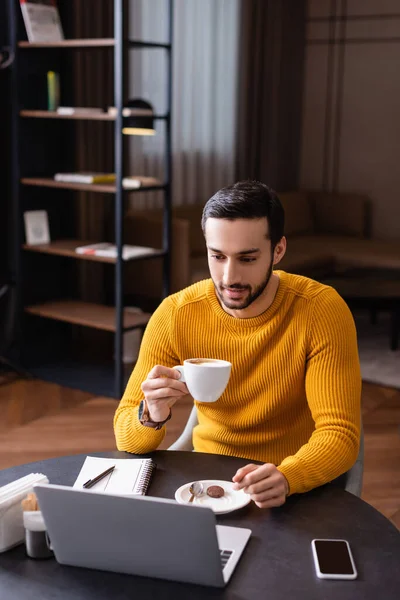 Молодий арабський фрілансер тримає чашку кави, дивлячись на ноутбук в ресторані — стокове фото