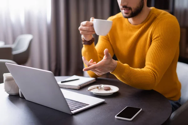 Обрізаний вид арабського фрілансера, який вказує на ноутбук, тримаючи чашку кави — стокове фото
