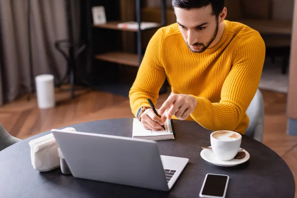 Arabian freelancer pointing with finger at laptop while writing in notebook in restaurant — Stock Photo