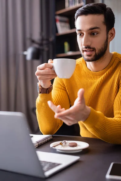 Изумленный арабский фрилансер, указывающий на ноутбук, держа чашку кофе в ресторане, размытый передний план — стоковое фото