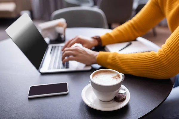 Vista recortada del freelancer escribiendo en el ordenador portátil cerca de la taza de café, fondo borroso - foto de stock