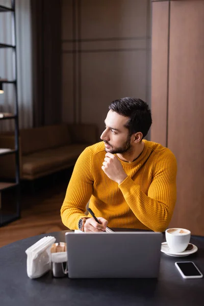 Pensive arabian freelancer holding pen near laptop and looking away in restaurant — Stock Photo