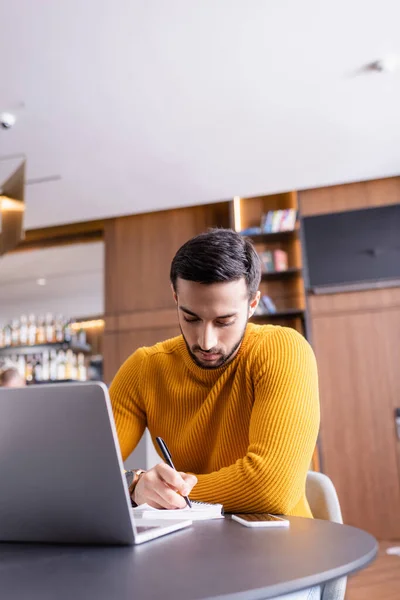 Joven árabe freelancer escribir en un cuaderno cerca de la computadora portátil y teléfono inteligente en el restaurante - foto de stock