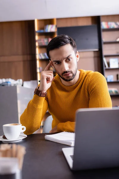 Thoughtful arabian freelancer looking at laptop while working in restaurant, blurred foreground — Stock Photo