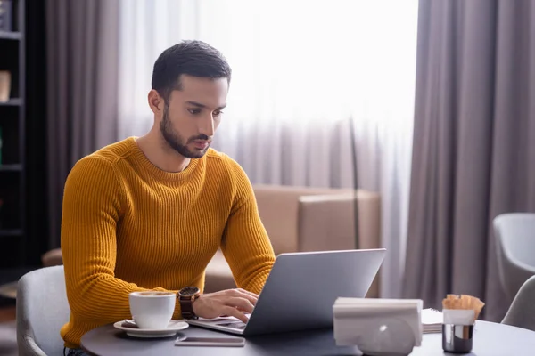 Teletrabajador árabe concentrado escribiendo en el ordenador portátil cerca de taza de café en el restaurante - foto de stock