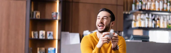 Excité homme arabe riant tout en tenant une tasse de café et regardant loin dans le restaurant, bannière — Photo de stock