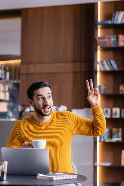 Feliz árabe freelancer mostrando gesto de victoria mientras sostiene la taza de café en el restaurante - foto de stock