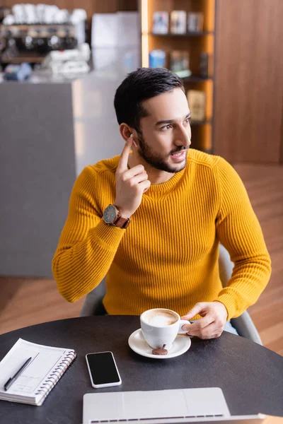 Молодий арабський фрілансер фіксує навушники, сидячи біля чашки кави та ноутбука — стокове фото