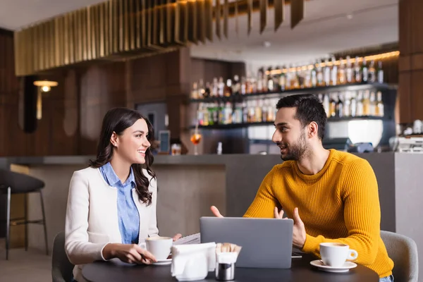 Joven árabe freelancer hablando con alegre amigo cerca de la computadora portátil y tazas de café en el restaurante, borrosa primer plano - foto de stock