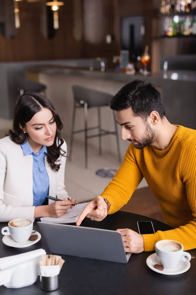 Arabian freelancer pointing at laptop near student writing in notebook in restaurant, blurred foreground — Stock Photo