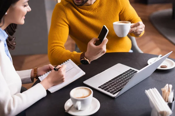 Vue recadrée de pigiste montrant smartphone à un ami écrit dans le carnet dans le restaurant — Photo de stock