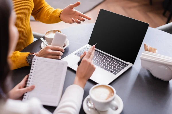 Man with smartphone pointing at laptop wit blank screen near coffee and friend with notebook on blurred foreground in restaurant — Stock Photo