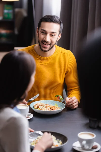 Homem árabe alegre segurando talheres perto de deliciosa salada, cappuccino e namorada em primeiro plano desfocado no restaurante — Fotografia de Stock